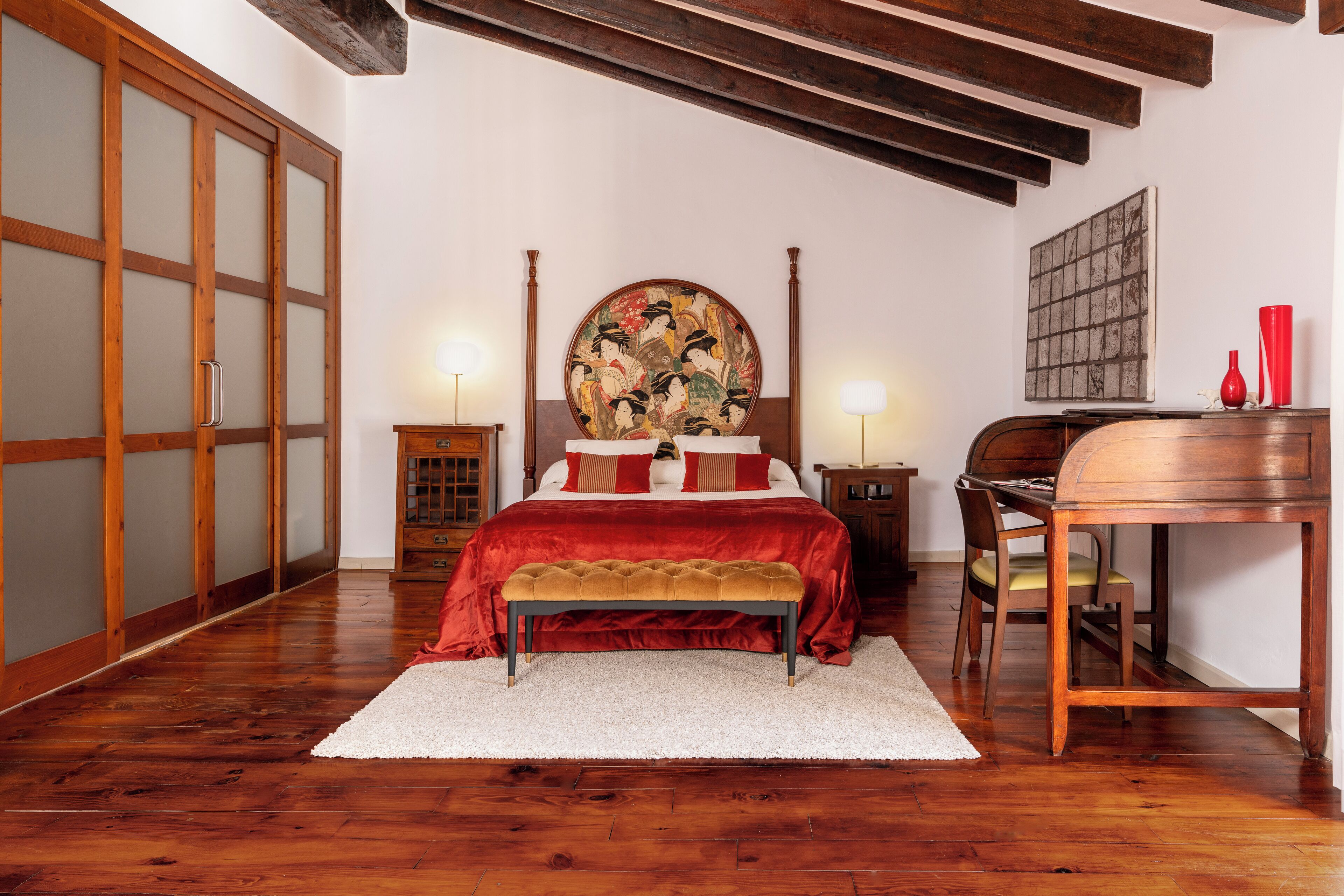Un dormitorio con suelo de madera, una cama con colcha roja, un cuadro redondo sobre la cama y un escritorio con una silla.