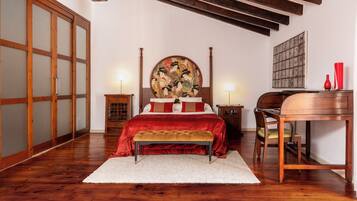 Un dormitorio con suelo de madera, una cama con colcha roja, un cuadro redondo sobre la cama y un escritorio con una silla.