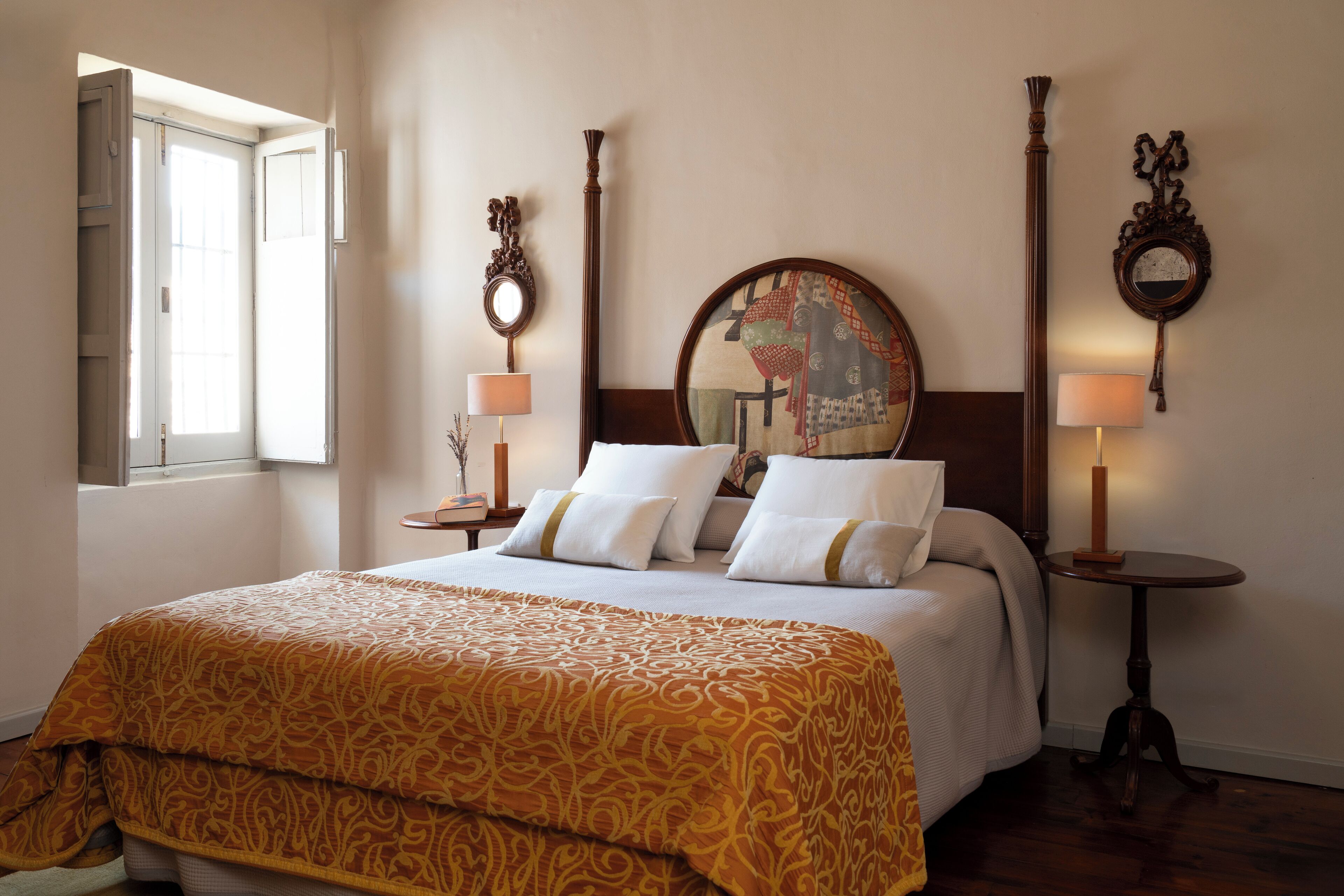 A hotel room with a large bed, two bedside tables, a round mirror, and a window with curtains.