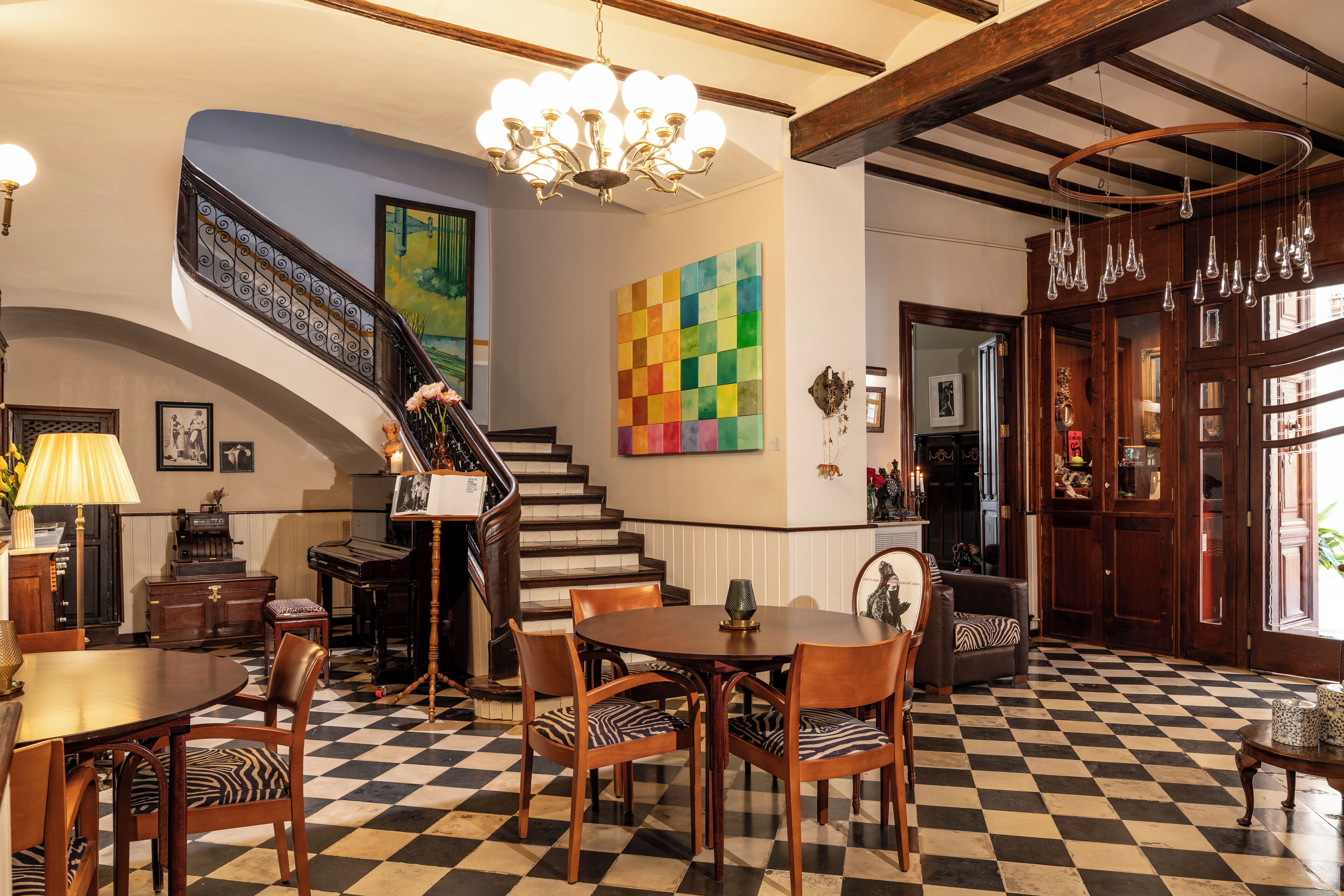 A checkered floor, a staircase, a dining area with chairs, a chandelier, and a colorful wall art piece.