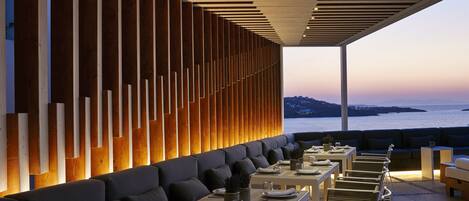 A modern outdoor dining area with a row of tables and benches, a wooden slatted wall, and a view of the sea.