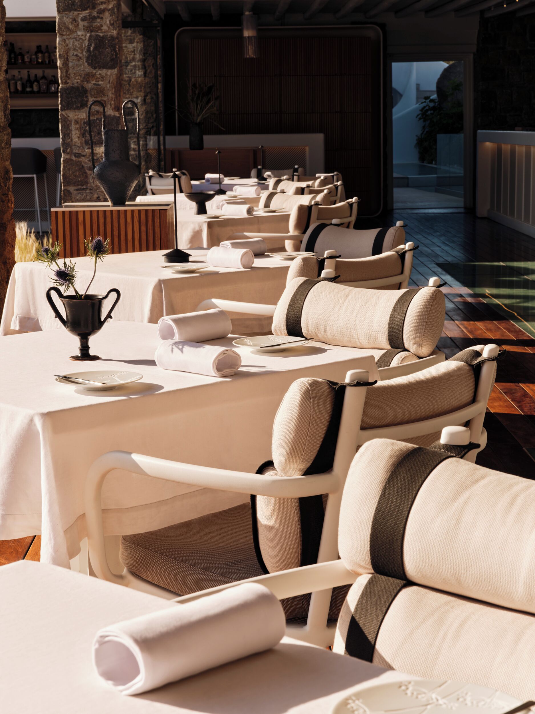 A restaurant with white tables and chairs, a black vase with flowers, and a stone wall in the background.