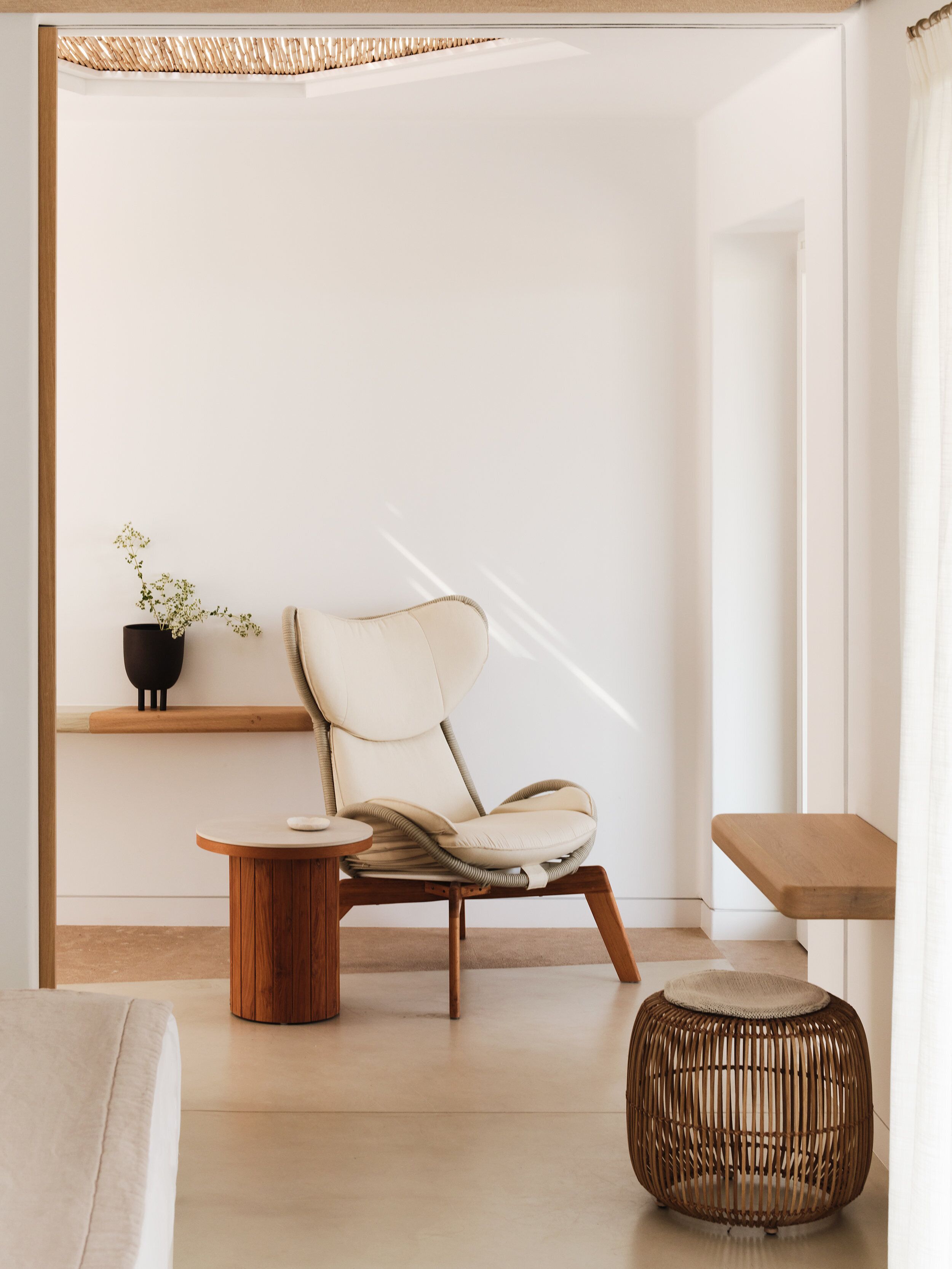 Una sala moderna con un sillón de color claro, una mesita redonda de madera, un otoman tejido y una pequeña planta en maceta sobre una estantería.