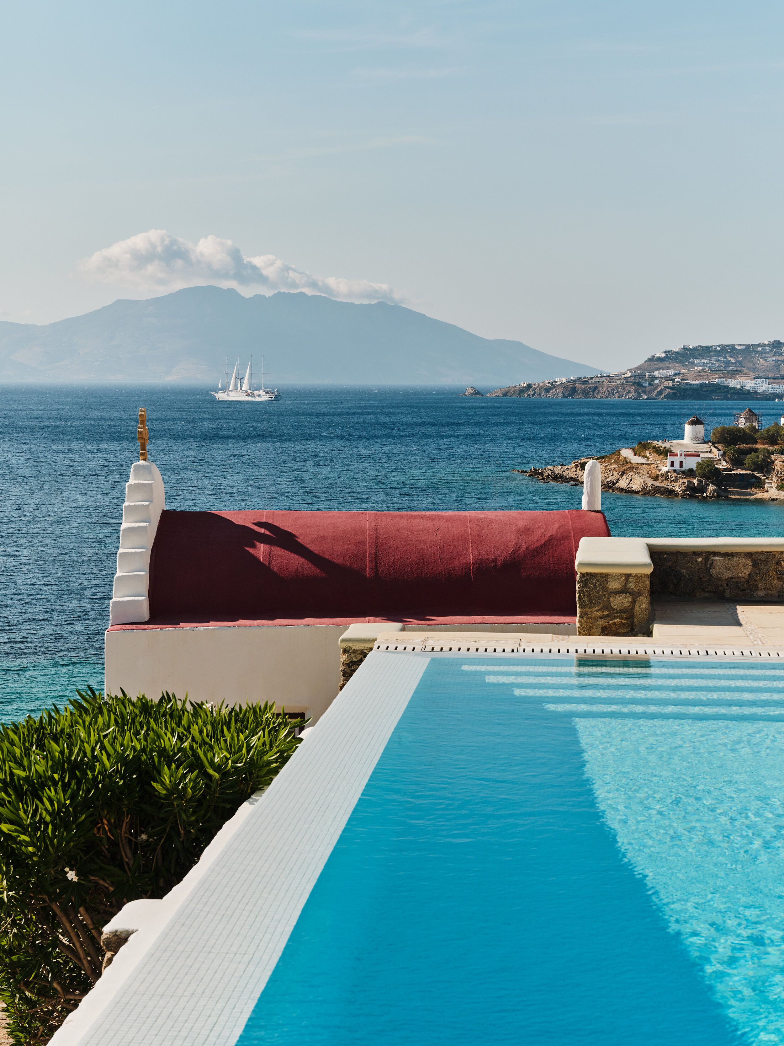 Vista de la piscina con una clara visión del mar, un velero y montañas distantes.