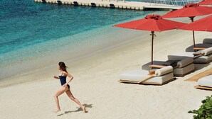 A beach with lounge chairs and umbrellas, a woman in a swimsuit walking, and clear blue water. - Bill&Coo Mykonos (Mykonos)