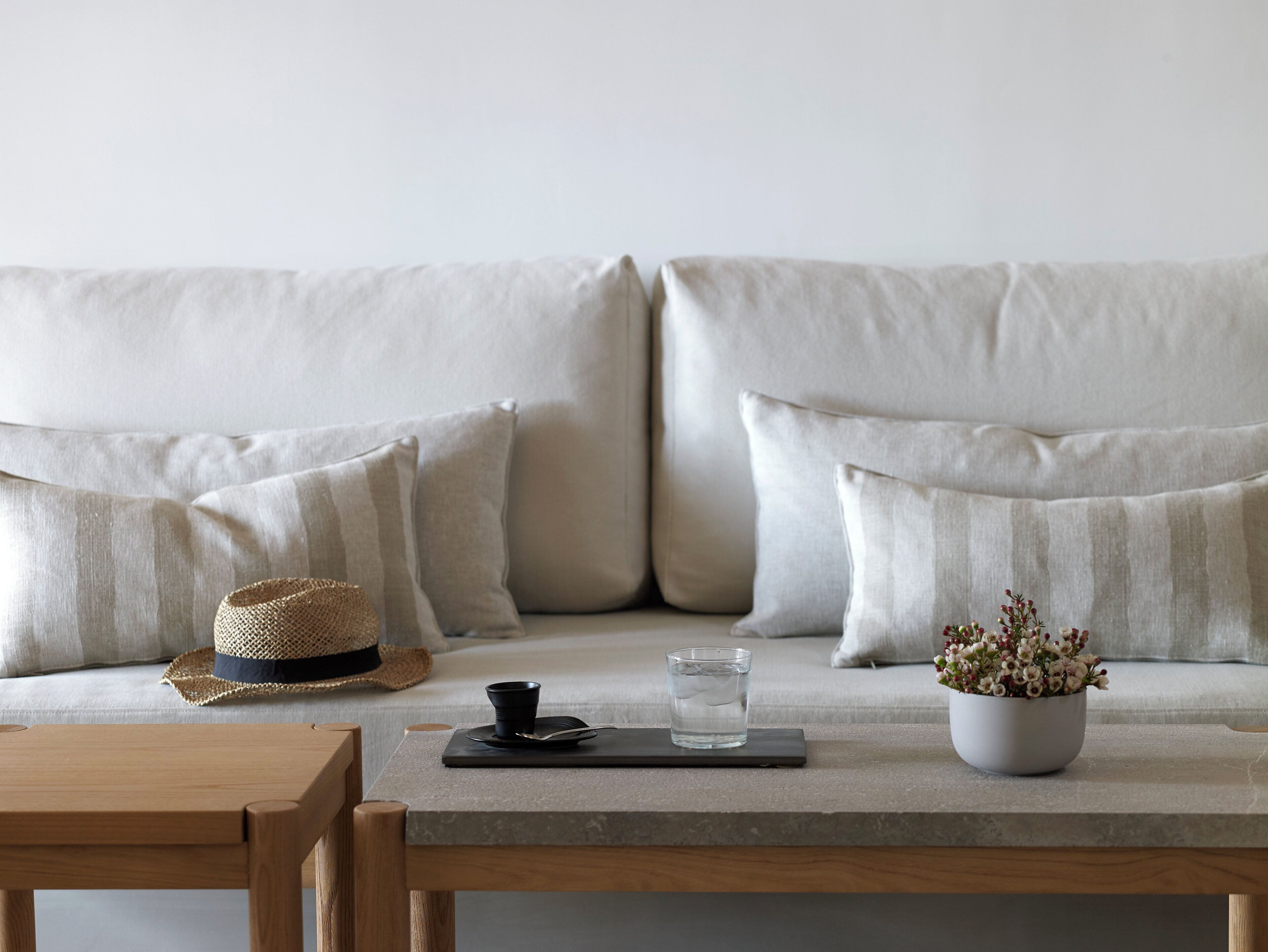 A modern living room with a beige sofa, a wooden coffee table, a small black tray with a cup and spoon, a glass of water, and a potted plant.