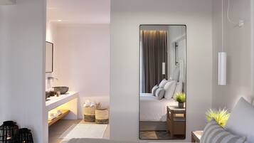 Habitación de hotel moderna con una cama grande, mesita de noche y un espejo que refleja el espacio. Se aprecia un baño con lavamanos, toallero y un pequeño estante con artículos de aseo.