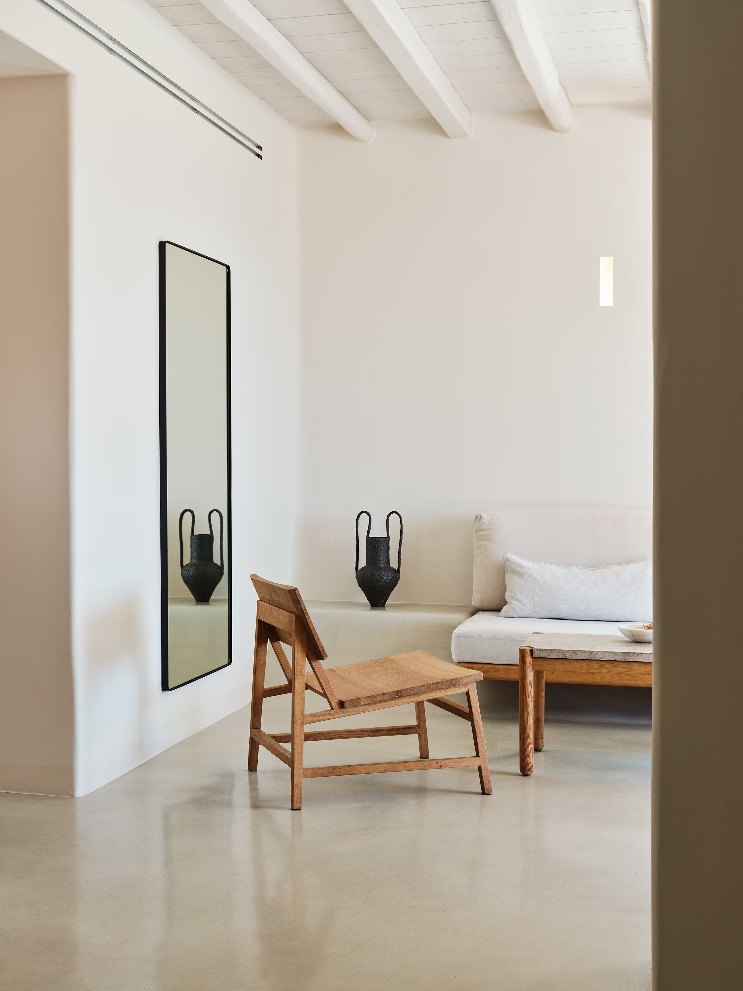 Een minimalistisch ingerichte kamer met een houten stoel, een bank en een grote spiegel aan de muur.
