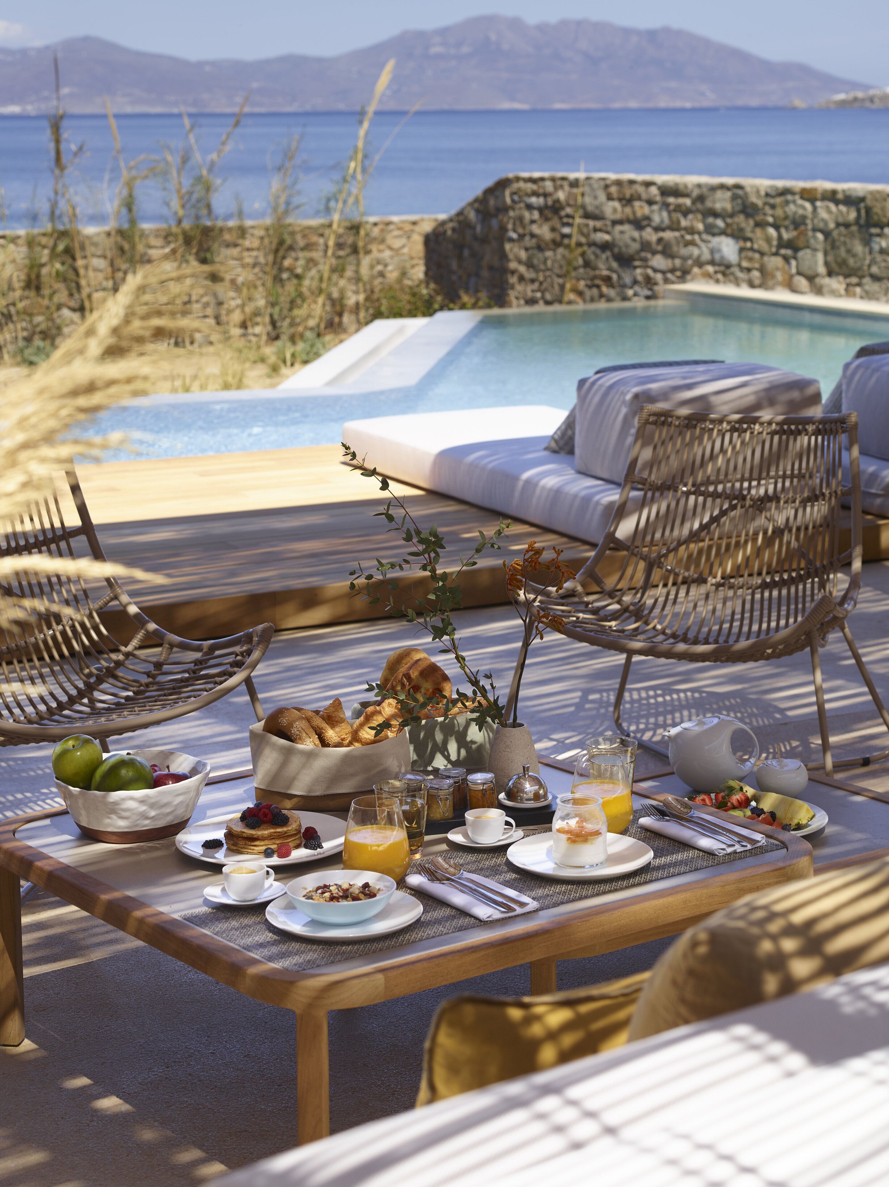Mesa de comedor dispuesta con alimentos para el desayuno, vista a una piscina y una playa, y sillas de mimbre.