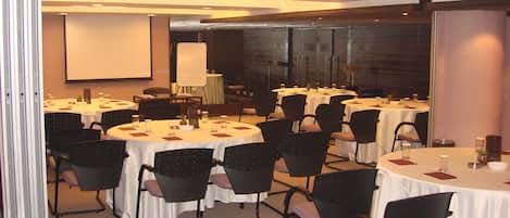 Un salón de conferencias con varias mesas redondas, sillas negras y una pantalla de proyector.
