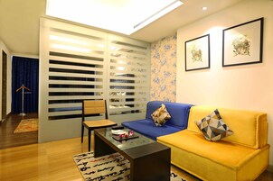 Ein Wohnzimmer mit einem blauen Sofa, einem gelben Sofa und einem Glastisch. Im Hintergrund befindet sich eine weiße Trennwand mit horizontalen Lamellen und ein blauer Vorhang.