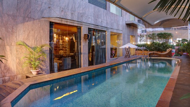 A modern indoor pool area with a clear pool, seating by the pool, and a potted plant. - Mosaic Original, Noida (Noida)
