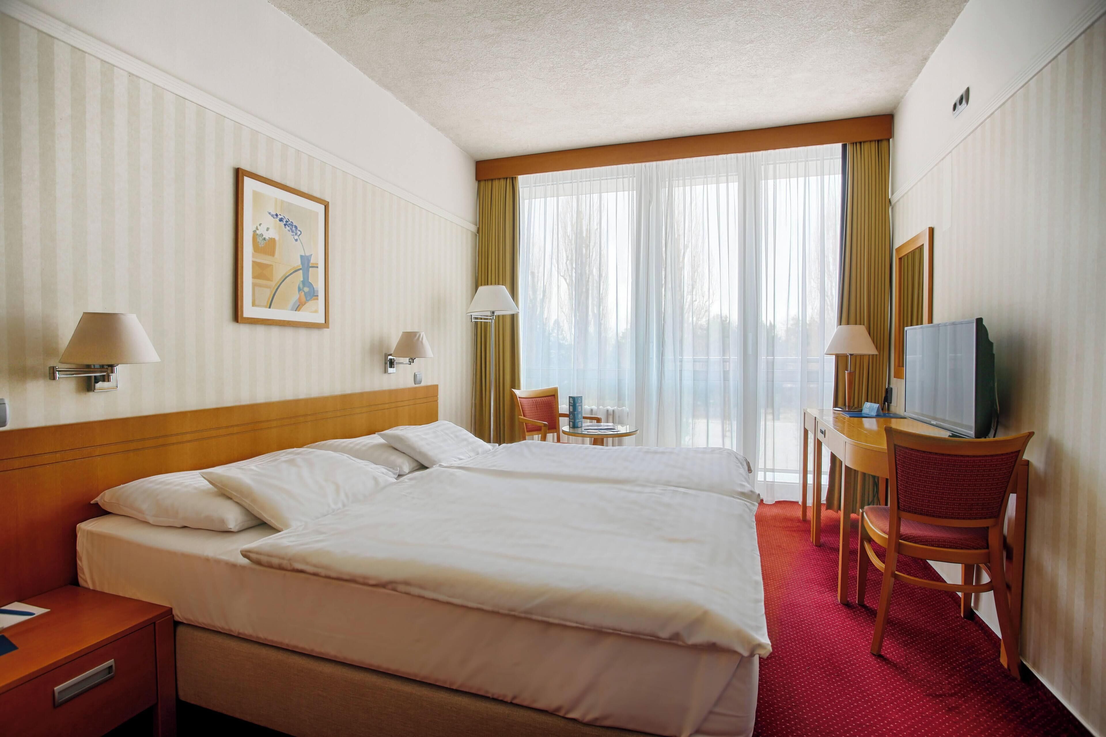 Habitación de hotel con una cama grande, dos mesitas de noche, un escritorio con silla, un televisor y una ventana con cortinas.
