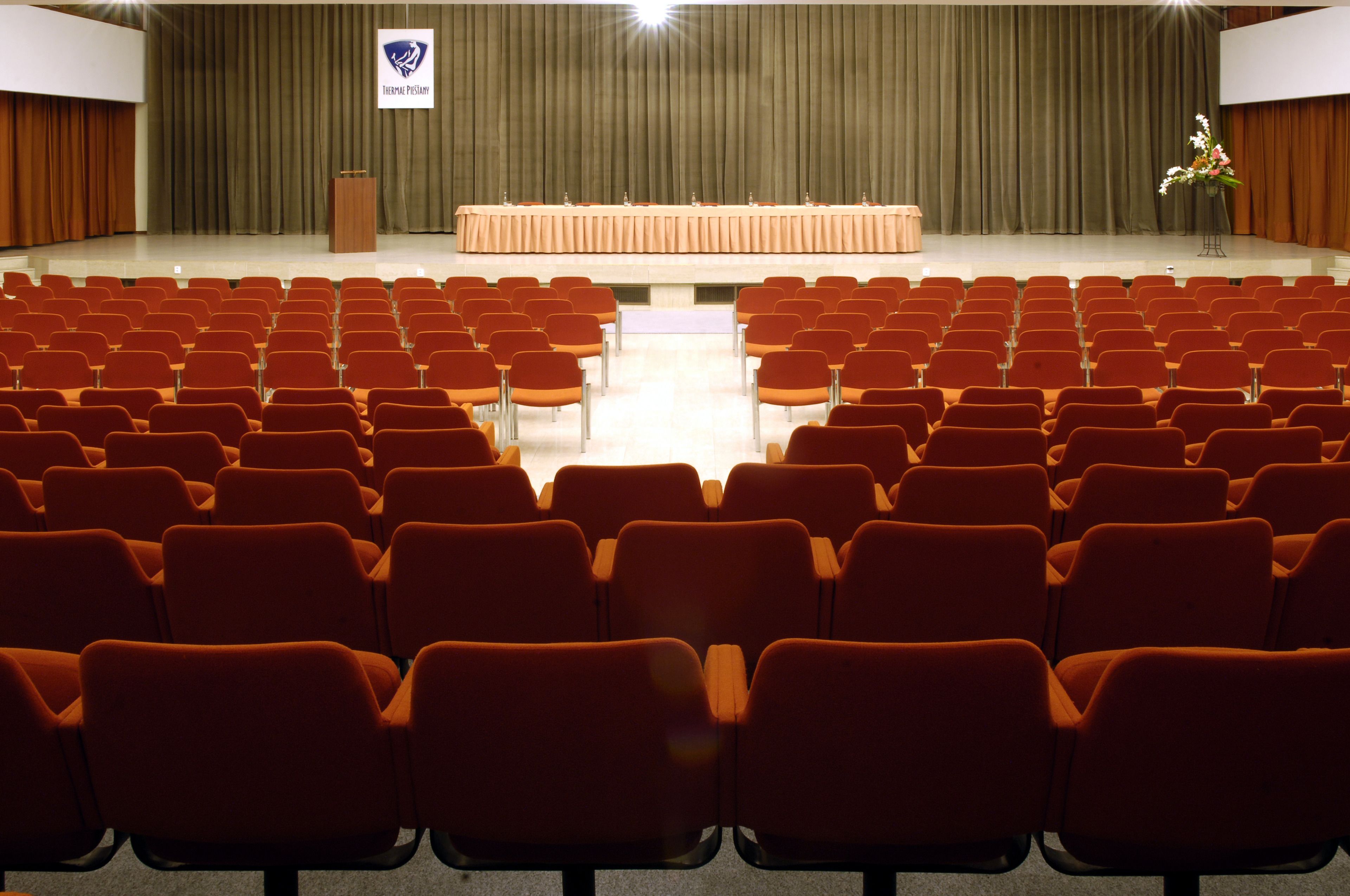 A conference room with rows of orange chairs, a podium, and a stage with a curtain backdrop.