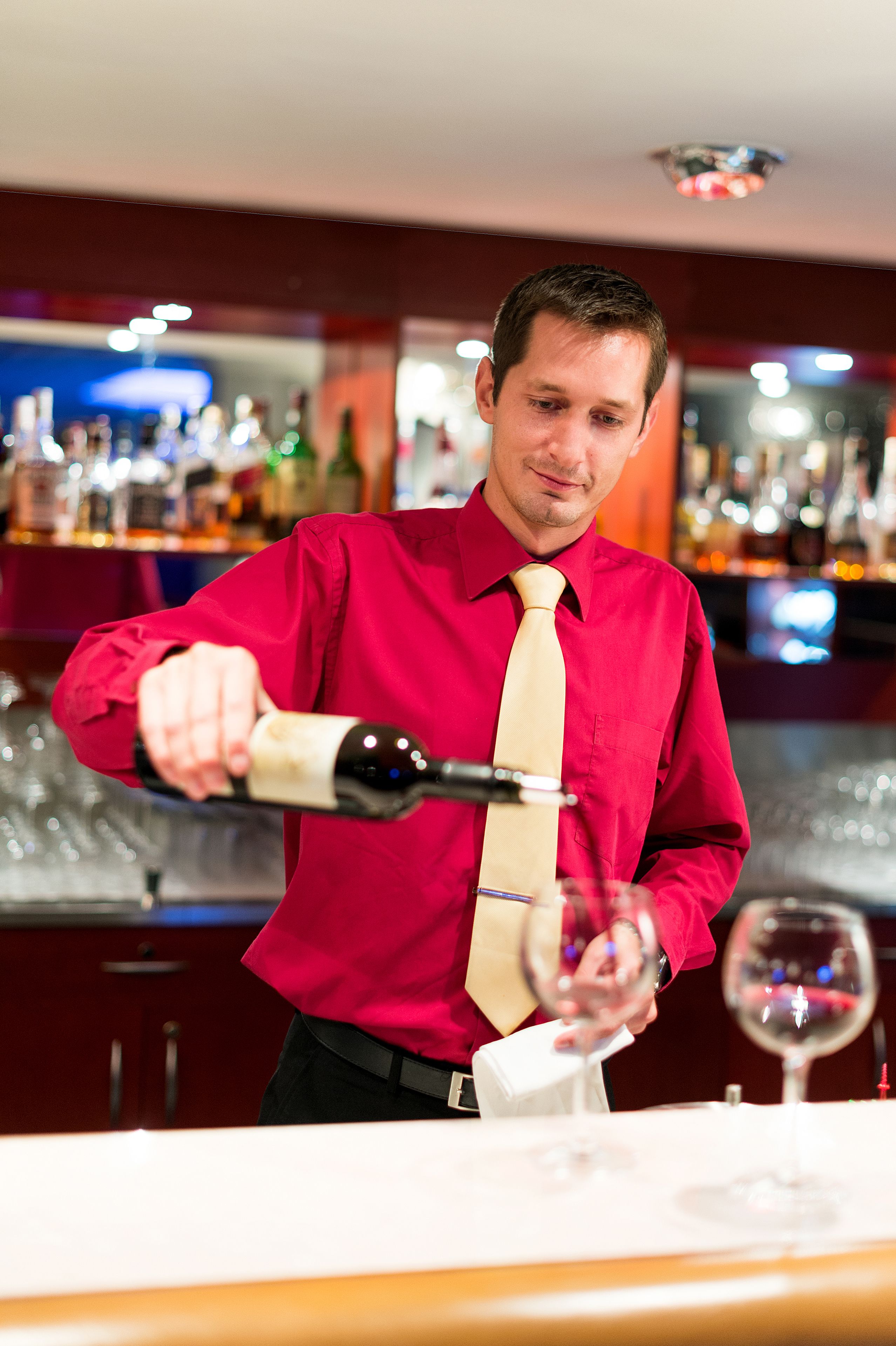 A person in a red shirt and beige tie pouring wine into a glass.