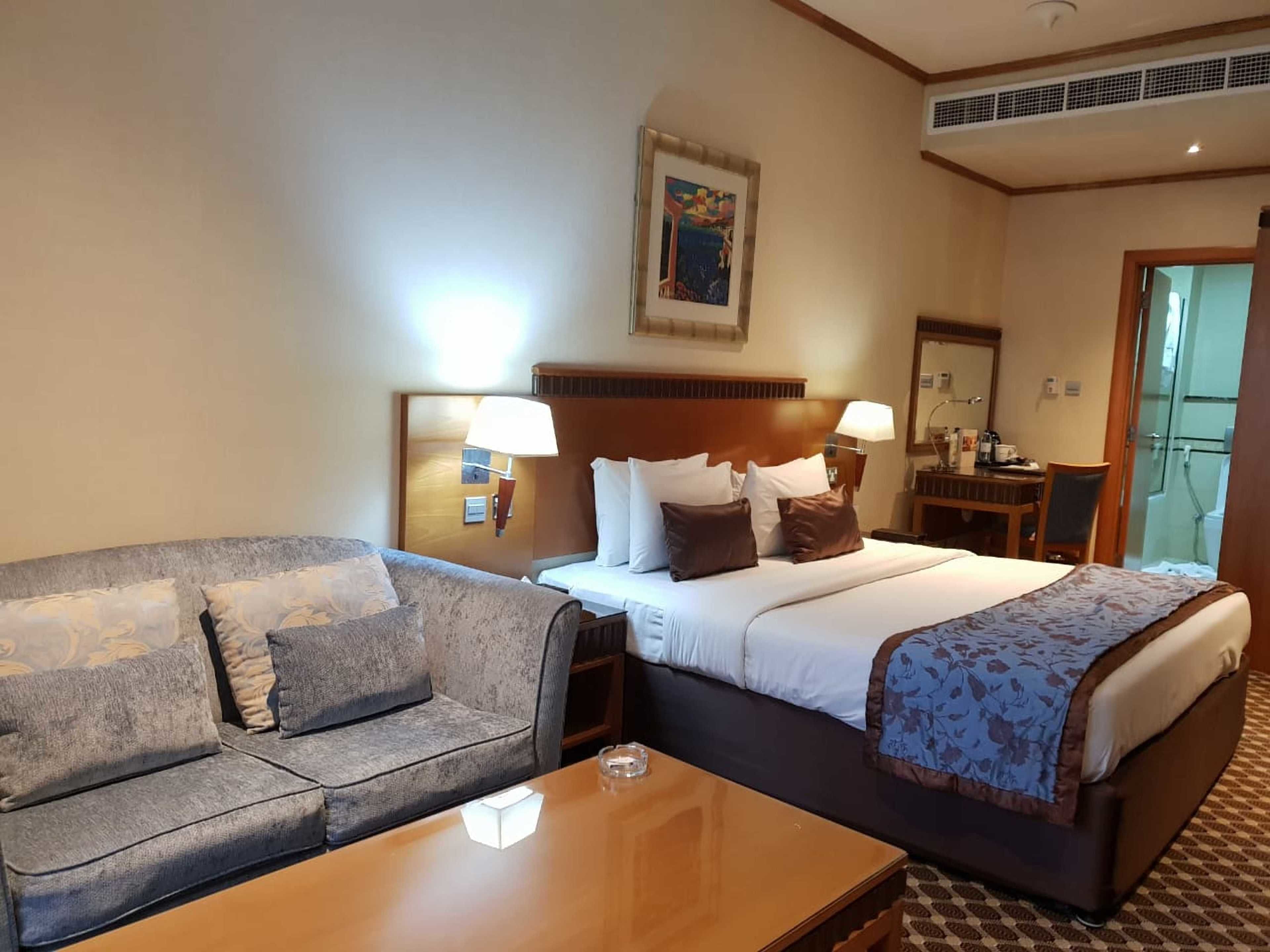 A hotel room with a bed, a sofa, a bedside table, a lamp, and a painting on the wall.