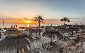 A beach with thatched umbrellas and lounge chairs, a palm tree, and the sunset over the ocean. - El Mouradi Palm Marina (Port El Kantaoui)