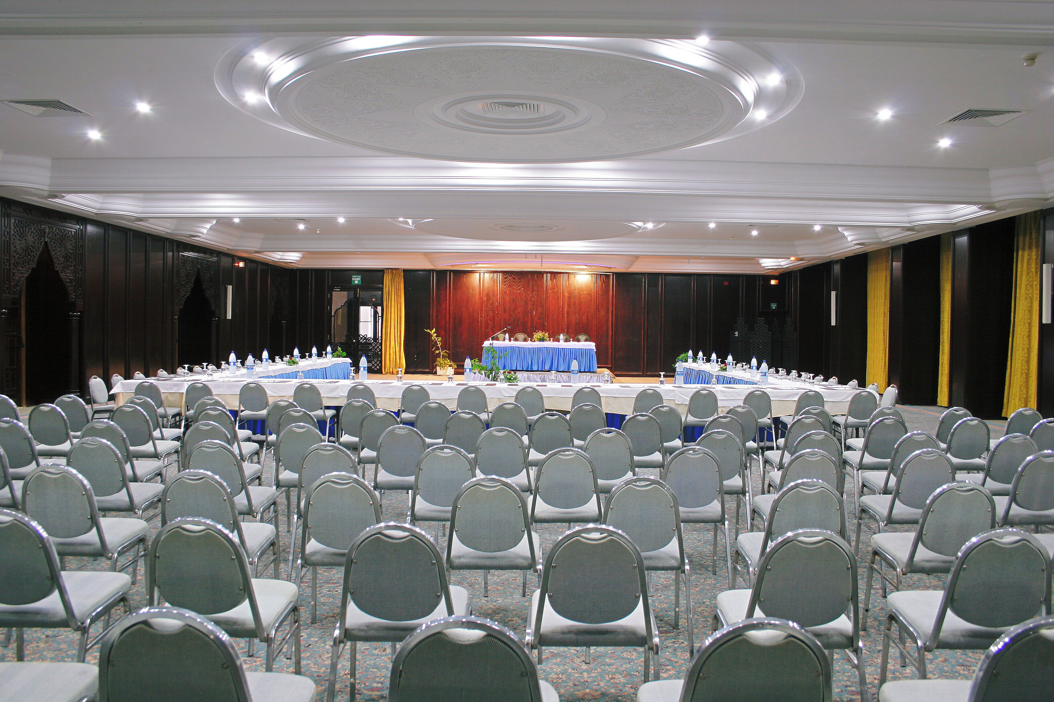 Un salón de conferencias con filas de sillas mirando hacia un escenario, una mesa azul con mantel blanco y una alfombra estampada.