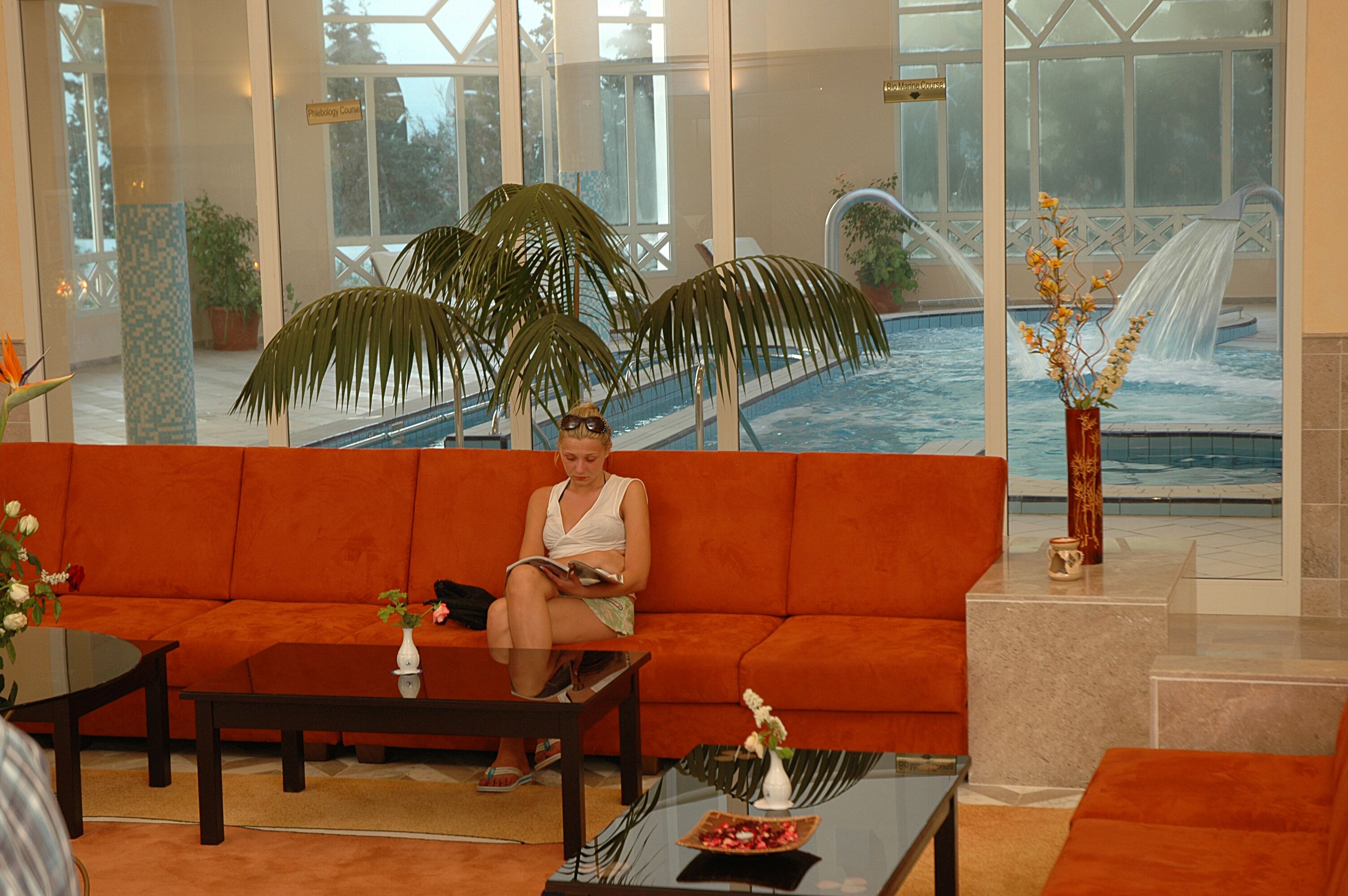 A woman sitting on an orange sofa in a well-lit lobby with a large window overlooking a pool area.