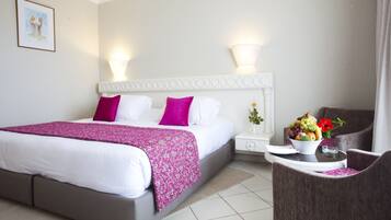 A hotel room with a large bed, a side table with a fruit bowl, a chair, and a painting on the wall.