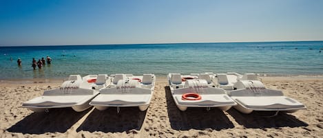 A beach with several white jet skis labeled "EL MOURADI" and "Club Restaurant" lined up on the sand, facing the sea.