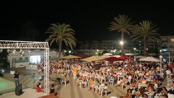 An outdoor seating area with numerous tables and chairs, palm trees, and a stage with a truss structure.