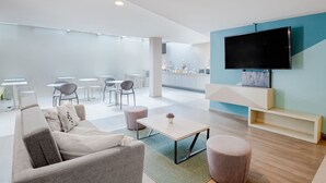 Una sala de estar moderna con un sofá, una mesa de centro y un reposapiés, un televisor de pantalla plana montado en la pared, y un comedor con sillas y mesas al fondo.