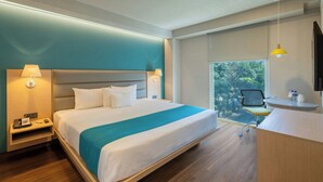 Una habitación de hotel moderna con una cama grande, lámparas de noche, un escritorio con una silla, una ventana con vista a árboles y un televisor montado.