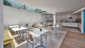 Un comedor moderno con mesas y sillas, una cocina al fondo y un tragaluz.