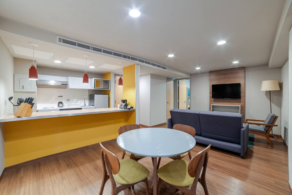Una cocina moderna con una isla central, un comedor con una mesa redonda y sillas, un sofá y un televisor de pantalla plana montado en la pared.