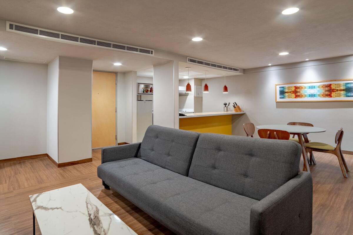 Una sala de estar moderna con un sofá gris, suelo de madera, una mesita de centro de mármol y un comedor con sillas de madera y una pequeña mesa.