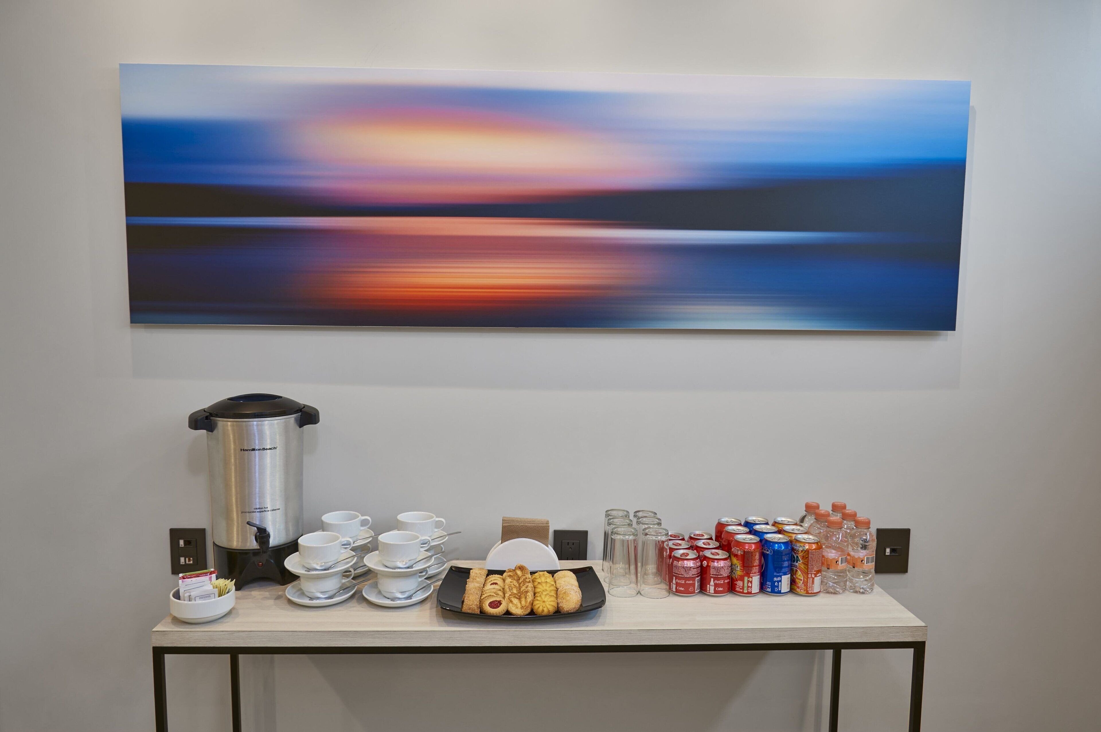 En frukostuppsättning med en kaffebryggare, koppar och bakverk på ett bord, och en tavla hänger ovanför.
