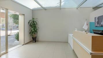 Zona de recepción con puerta de vidrio que da al exterior, mostrador de recepción, planta en maceta y un cartel en la pared.