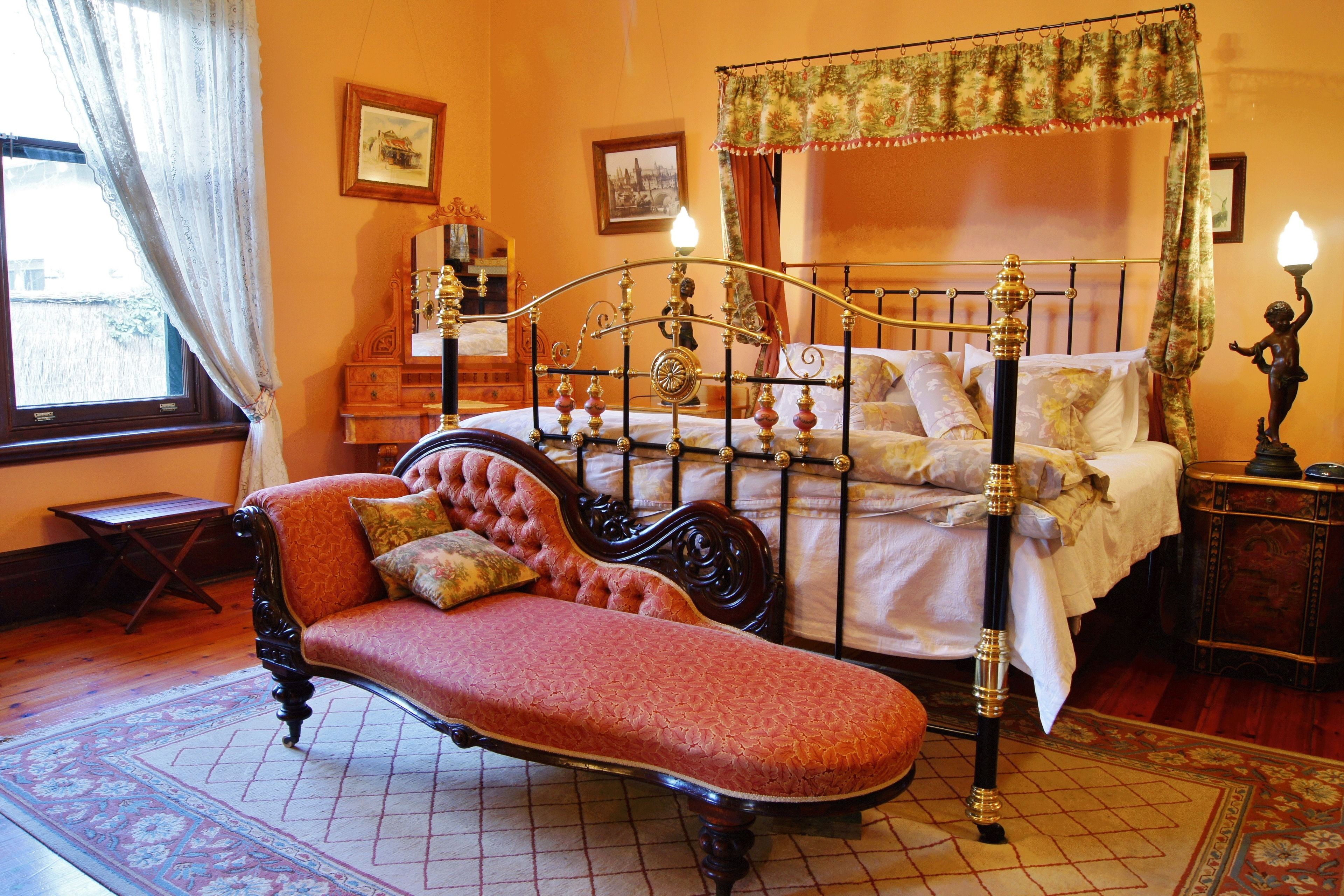 A traditional bedroom with a four-poster bed, a chaise lounge, and a nightstand with a lamp.