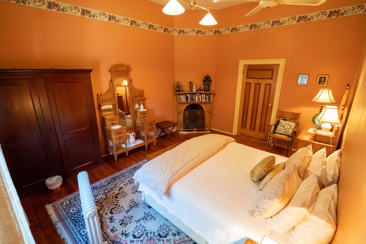 A bedroom with a large bed, a wooden wardrobe, a dresser, a mirror, a fireplace, and a nightstand with a lamp.