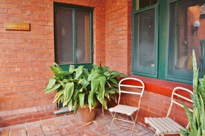 Un bâtiment en briques sur lequel on peut lire l’inscription « Jardin et lofts ». Devant le bâtiment, on trouve deux chaises en métal blanc et une grande plante en pot.