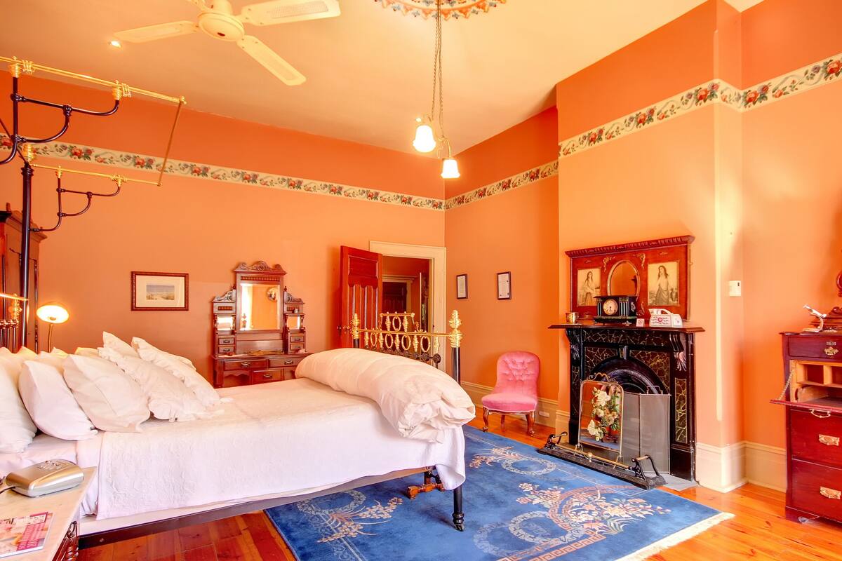 A bedroom with a bed, a dresser, a mirror, a fireplace, and a ceiling fan.