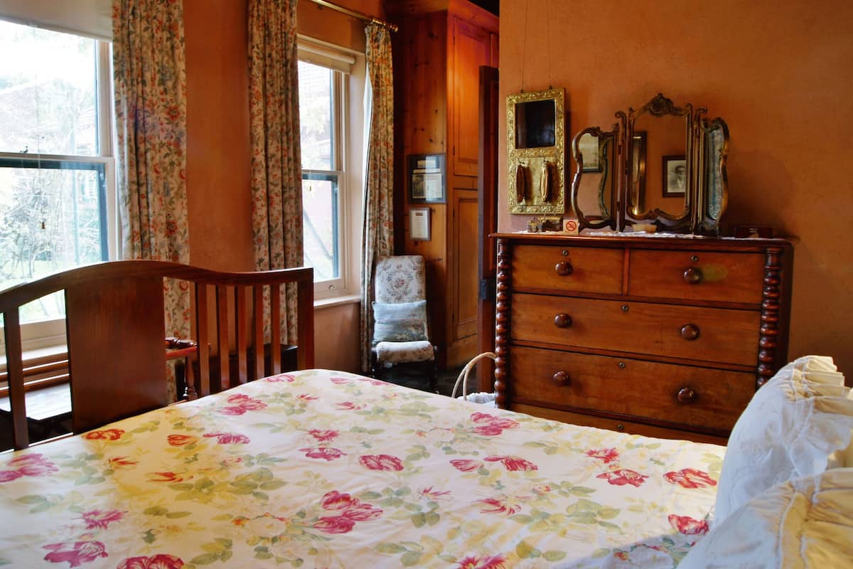 A bedroom with a floral bedspread, a wooden dresser with mirrors, a chair, and a window with curtains.