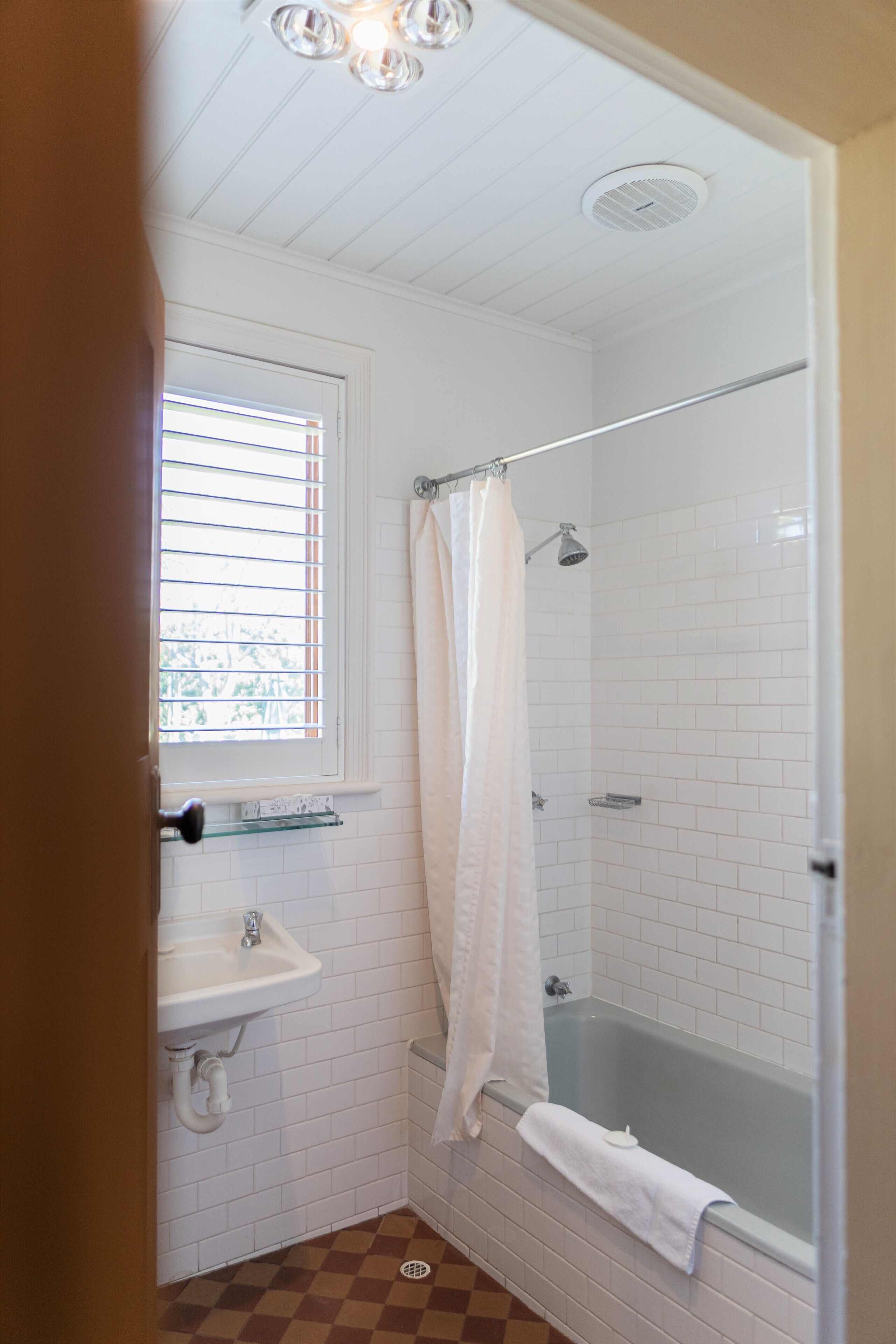 Une salle de bain avec un lavabo blanc, une fenêtre avec des stores, une douche avec une porte en verre et un carrelage à carreaux.