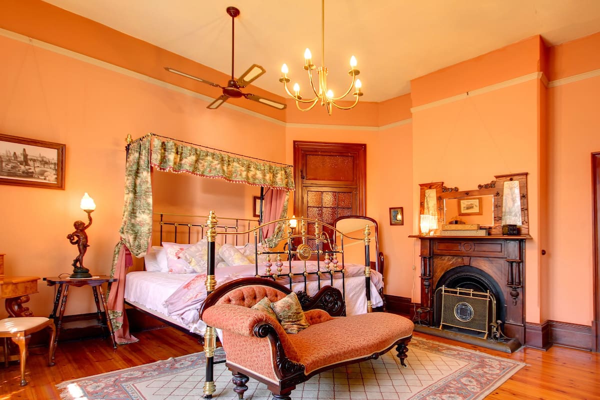 A bedroom with a four-poster bed, a chaise lounge, a fireplace, and a ceiling fan.
