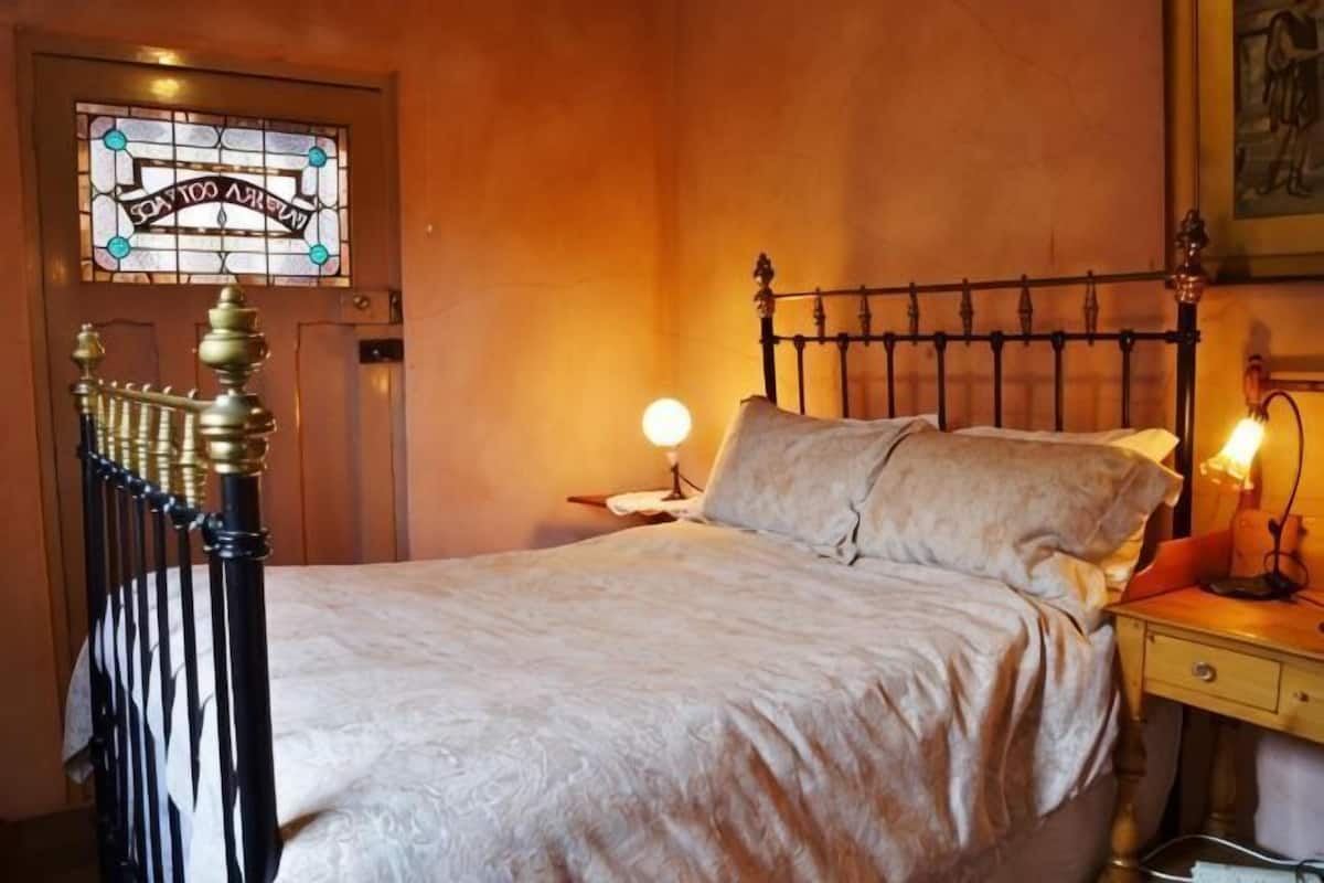 A bed with a headboard, a nightstand with a lamp, a wall-mounted clock, and a stained glass window.