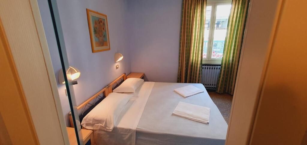 A hotel room with a bed, two bedside lamps, a window with curtains, and a painting on the wall.