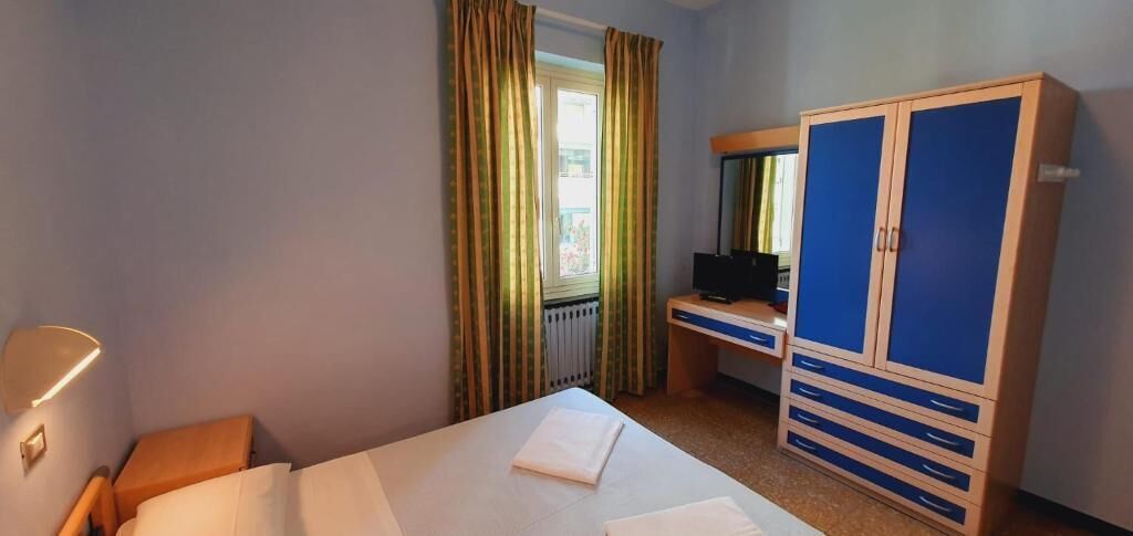 A hotel room with a bed, a desk with a computer, a wardrobe, and a window with curtains.