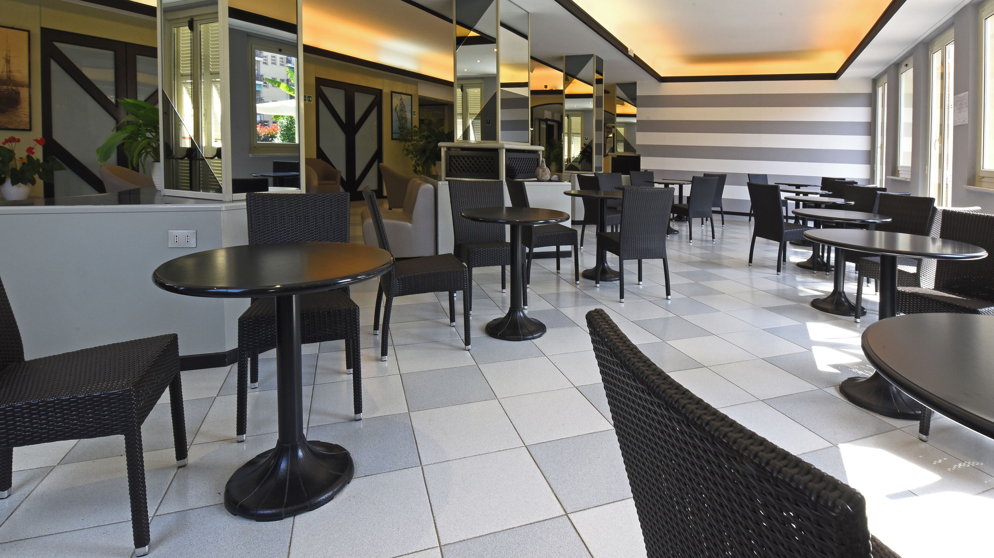 A modern cafe with black wicker furniture, white tiled floors, and large windows allowing natural light.