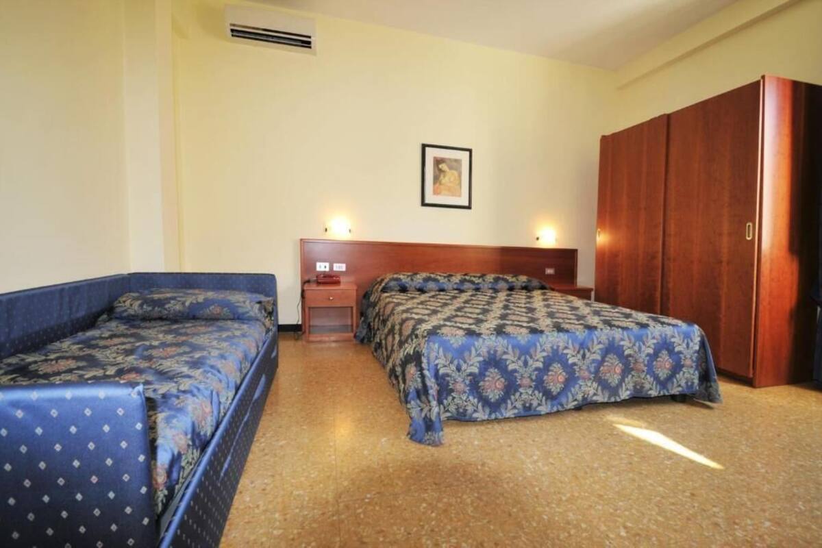 A hotel room with two beds, a wooden wardrobe, a nightstand, and a framed picture on the wall.