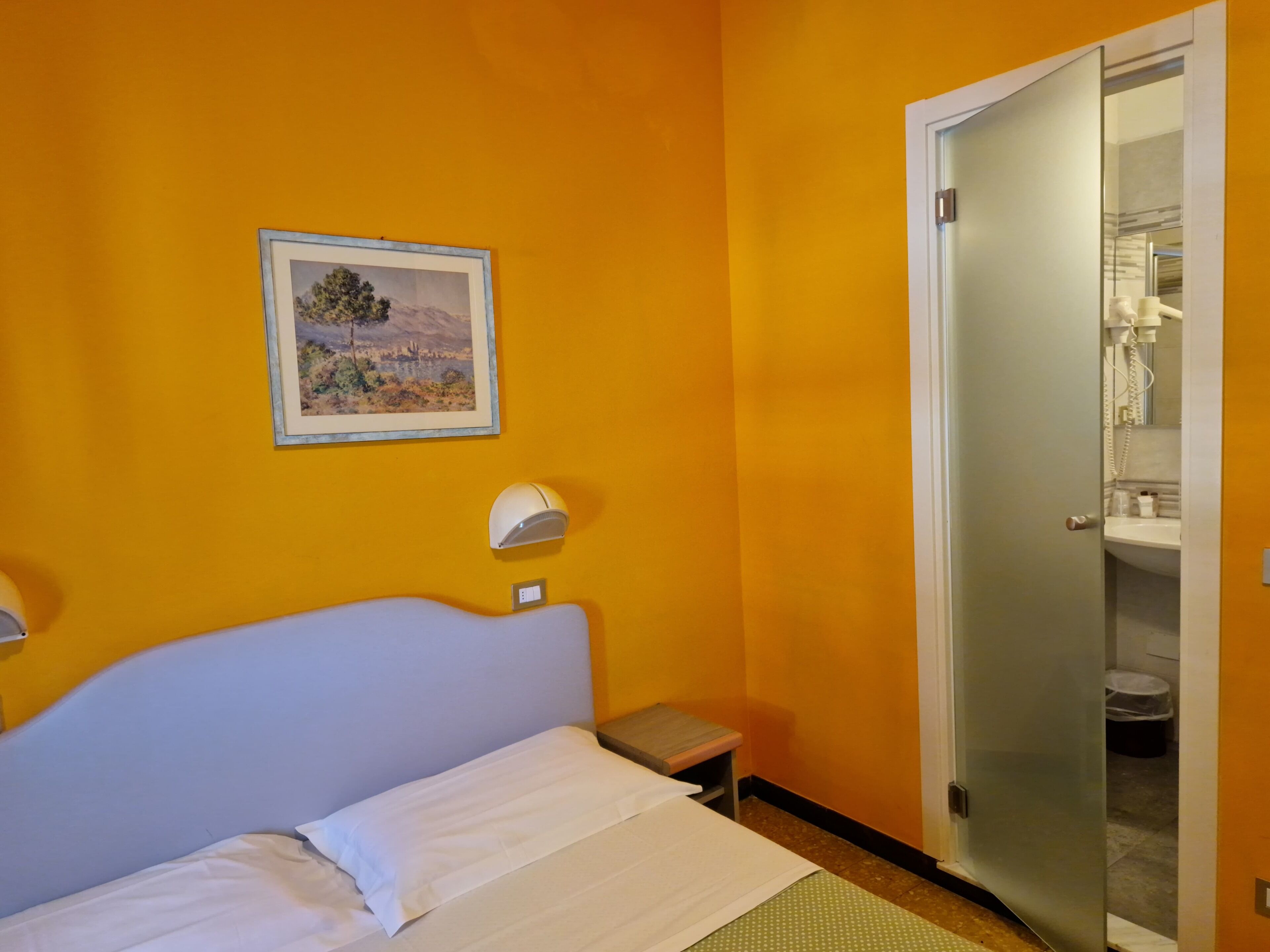 Una habitación con cama, una mesita de noche pequeña, una lámpara de pared, un cuadro enmarcado y una puerta que da al baño.