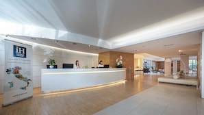 Ein moderner Hotel-Empfangsbereich mit einer weißen Rezeption, einem großen Poster, das das Hotel bewirbt, und einer geräumigen Lobby mit Holzboden und einer Sitzecke.