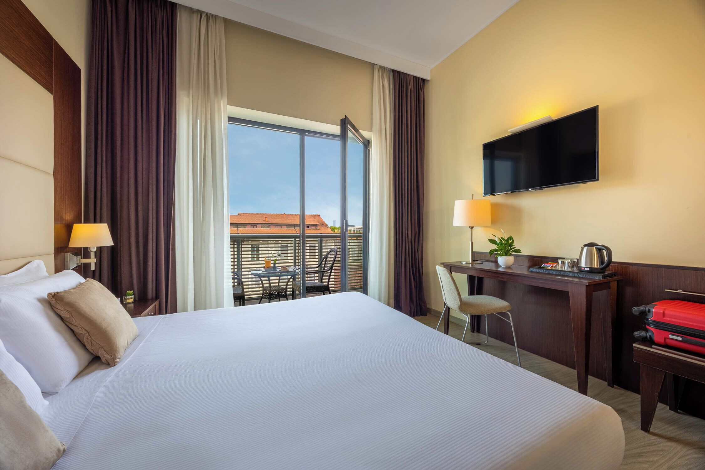 A hotel room with a large bed, a desk with a TV, a chair, a small table with a plant, and a window with curtains.