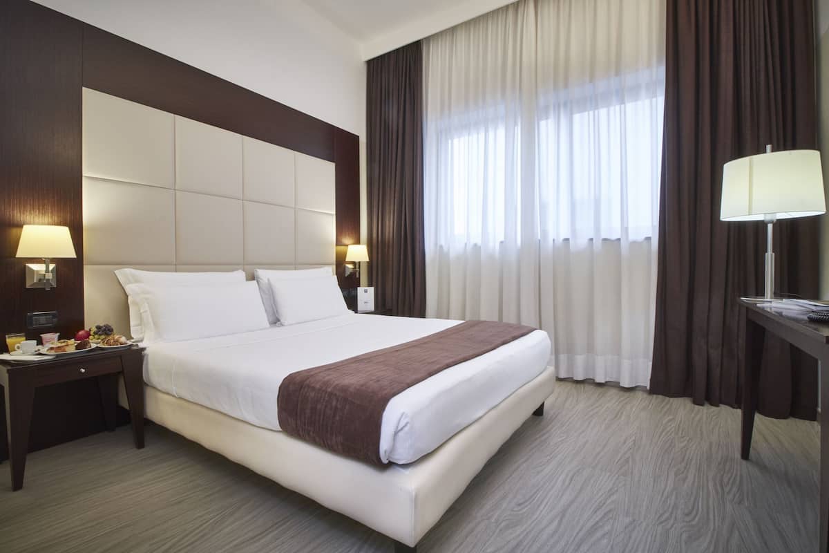 Ein Hotelzimmer mit einem großen Bett, zwei Nachttischen mit Lampen, einem zusätzlichen Tischchen mit einer Lampe und einem Fenster mit Vorhängen.