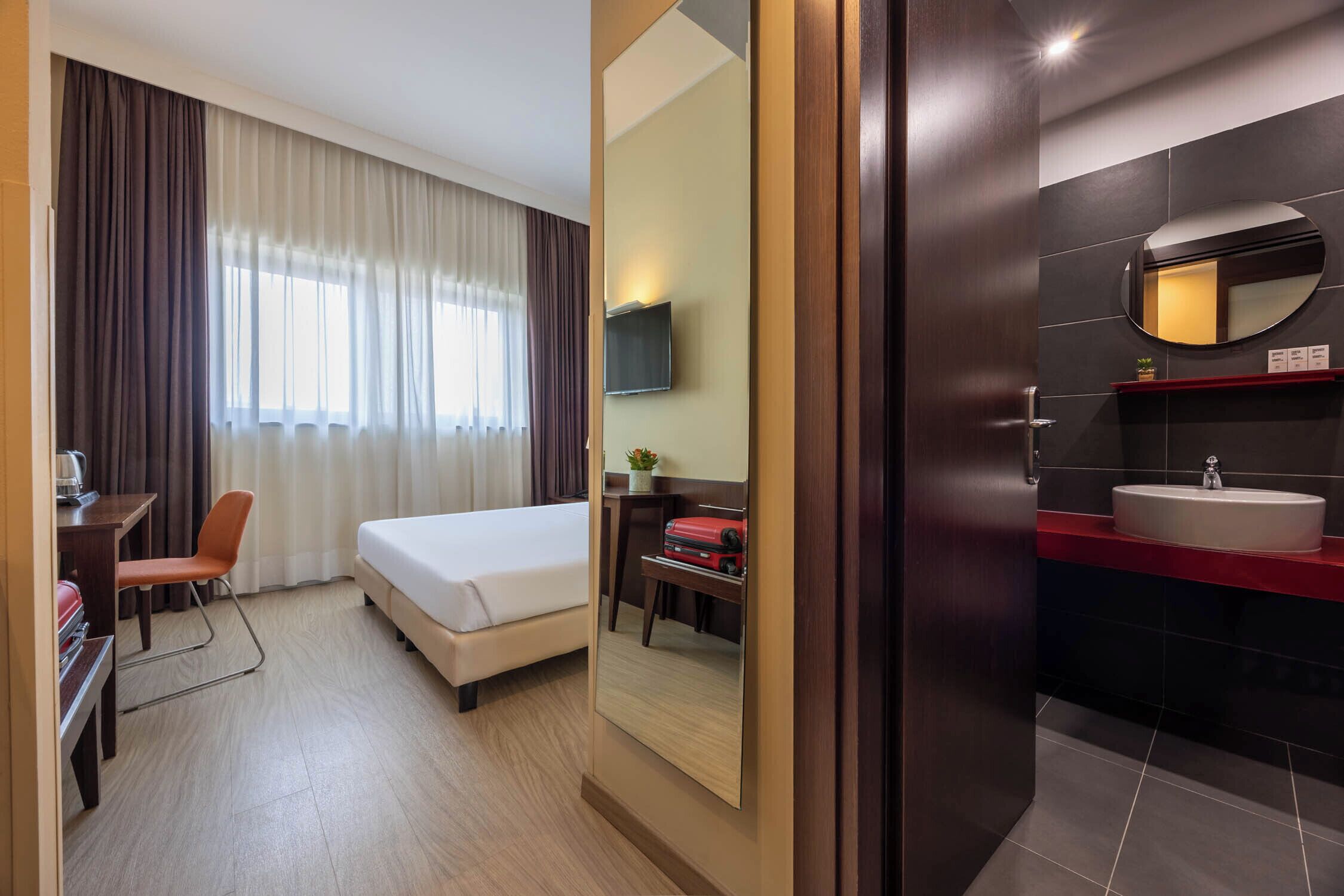 Hotelkamer met een bed, een bureau met stoel, een aan de muur bevestigde televisie, een badkamer met wastafel en spiegel, en een groot raam met gordijnen.